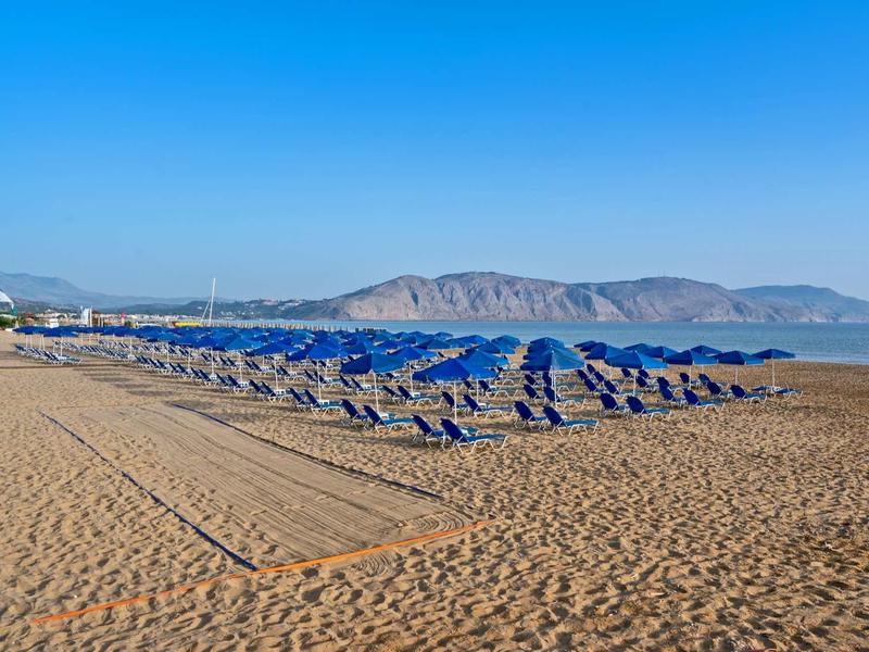 Breiter Sandstrand mit Reihen blauer Sonnenschirme und Bergen im Hintergrund unter klarem Himmel.