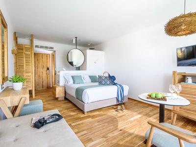 Chambre d'hôtel lumineuse avec lit double, meubles en bois et coin salon avec table et chaises.