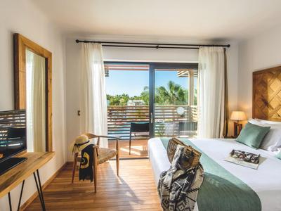 Chambre d'hôtel lumineuse avec grand lit, meubles en bois et balcon avec vue sur des palmiers.