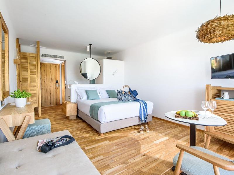 Chambre d'hôtel lumineuse avec lit double, meubles en bois et coin salon avec table et chaises.