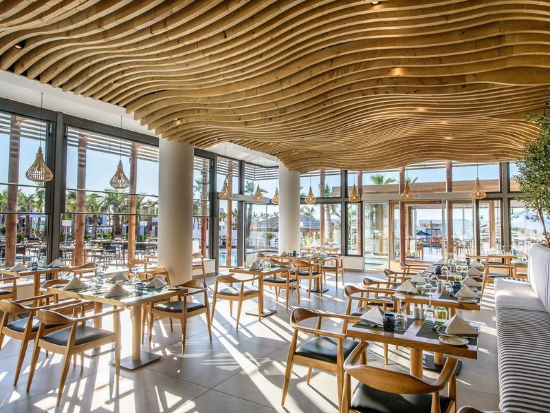 Restaurant moderne avec plafond ondulé, intérieur en bois clair, lumière naturelle abondante et grandes fenêtres.