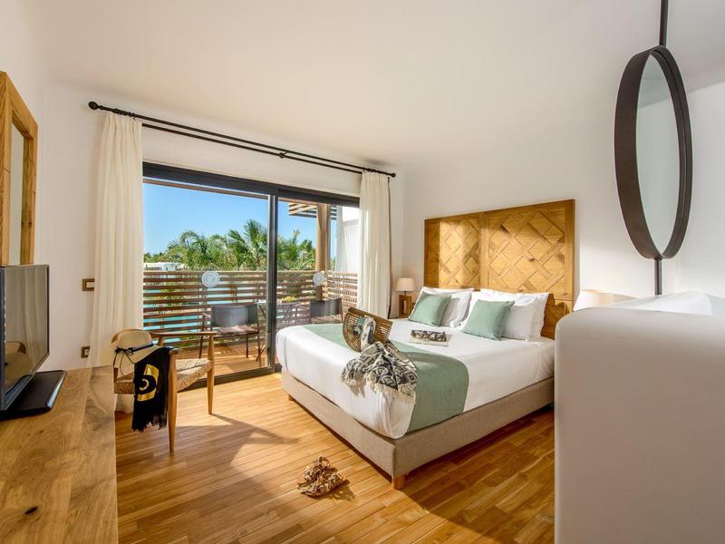 Chambre d'hôtel lumineuse avec lit double, meubles en bois et balcon avec vue sur les palmiers.