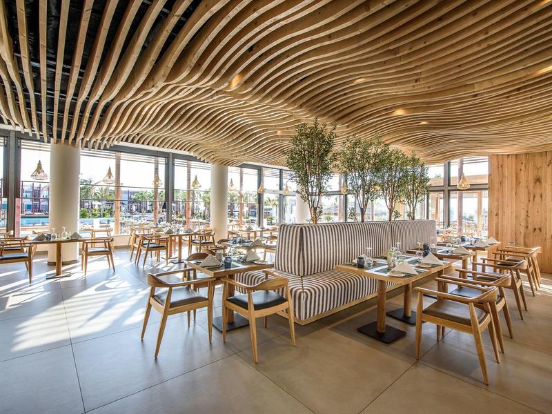 Restaurant lumineux avec plafond en bois, grandes fenêtres et tables et chaises élégantes.