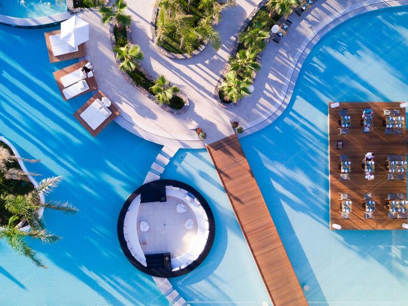 Vue aérienne d'un hôtel avec piscine, transats, pavillons et passerelle sur l'eau.