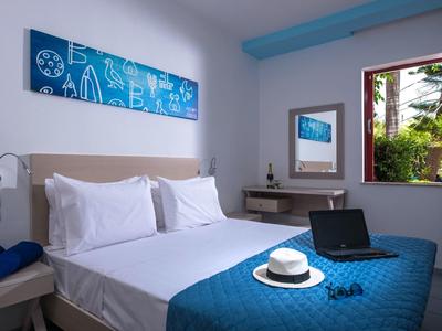 Lichte hotelkamer met tweepersoonsbed, blauw sprei, laptop en open raamzicht.