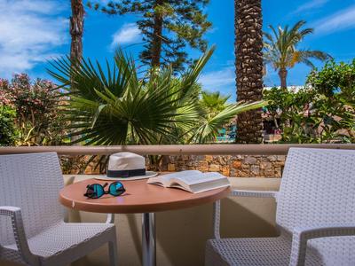 Balkon mit zwei weißen Stühlen, einem Tisch, einer offenen Zeitschrift und Sonnenbrille, umgeben von Palmen.