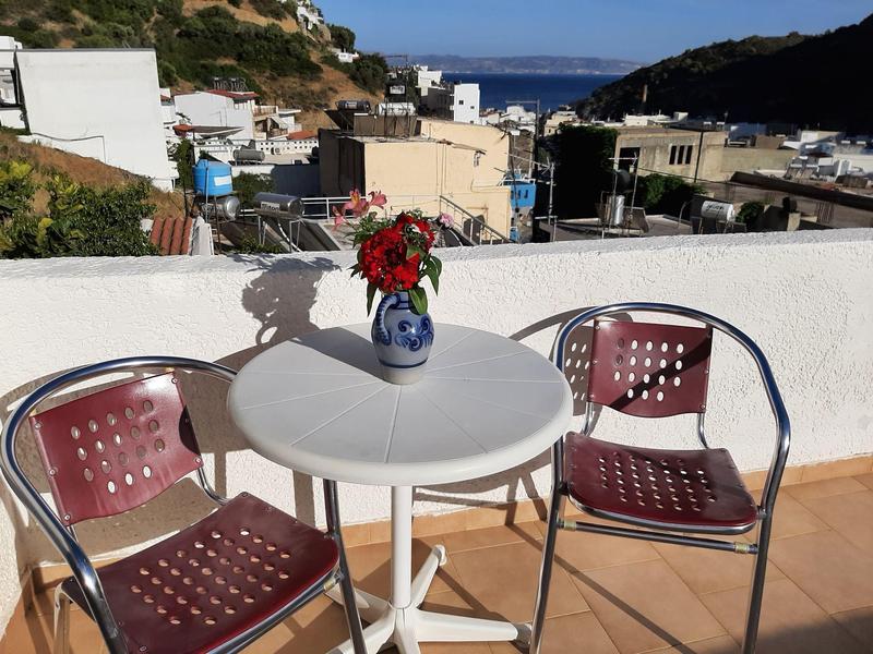 Kleiner runder Tisch mit Blumen auf einer Terrasse mit zwei Stühlen und Blick auf Häuser und Hügel.