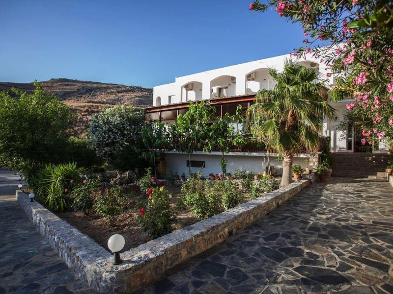 Weißes Gebäude mit Balkon, umgeben von üppigen Gärten und Steinweg unter blauem Himmel.