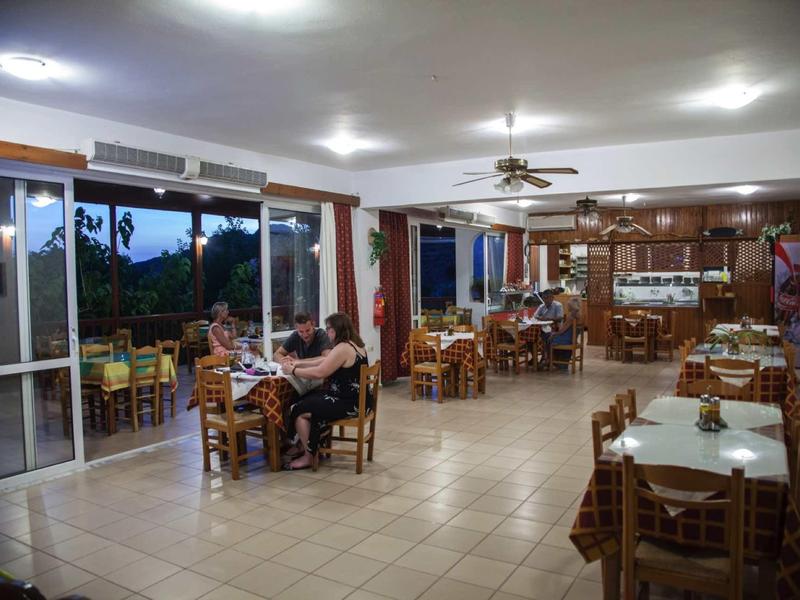 Ein großes Esszimmer mit mehreren Holztischen und Stühlen, beleuchtet durch Deckenlampen und große Fenster.