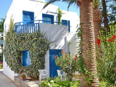 Bâtiment blanc avec fenêtres bleues et balcon entouré de palmiers et de fleurs.