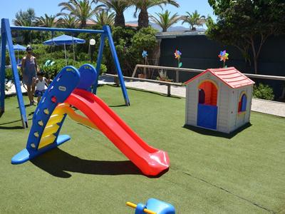 Parco giochi per bambini con scivolo rosso, struttura blu per arrampicata e casetta su prato artificiale verde.