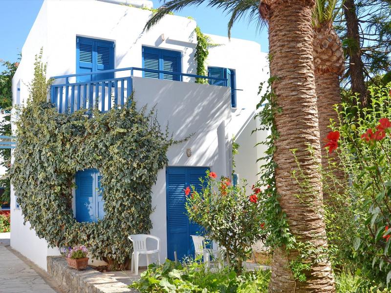 Casa bianca con persiane blu e balcone, circondata da piante verdi e palme.