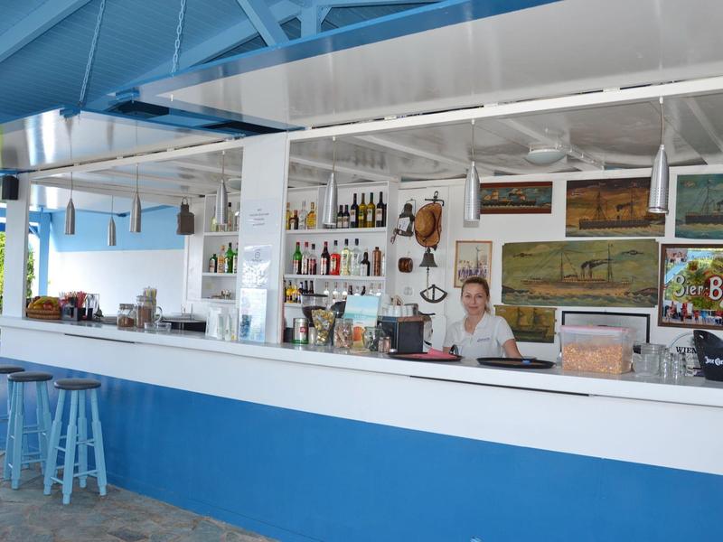 Bar sulla spiaggia aperta con bancone bianco e blu e un barista in un interno luminoso.