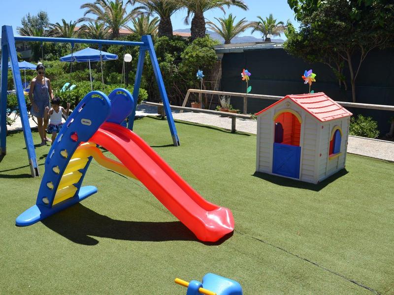 Parco giochi per bambini con scivolo rosso, struttura blu per arrampicata e casetta su prato artificiale verde.