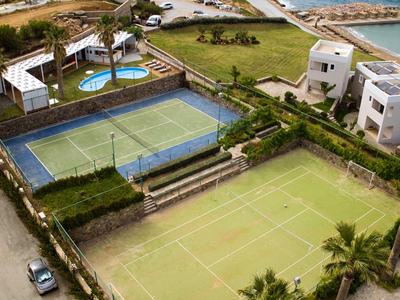 Luchtfoto van een resort met tennisbanen, zwembaden en witte gebouwen aan het water.