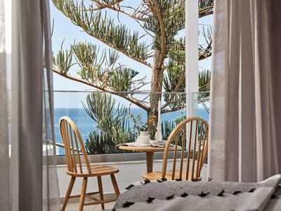 Blick durch geöffnete Glasfenster auf Balkon mit Rundtisch, zwei Holzstühlen und Meereskulisse.