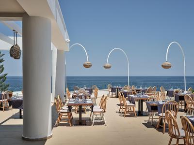 Openluchtrestaurant met uitzicht op zee, gedekte tafels en houten meubels onder een blauwe lucht.