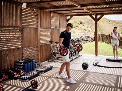 Persoon traint met een medicijnbal in een open houten paviljoen met fitnessapparatuur.