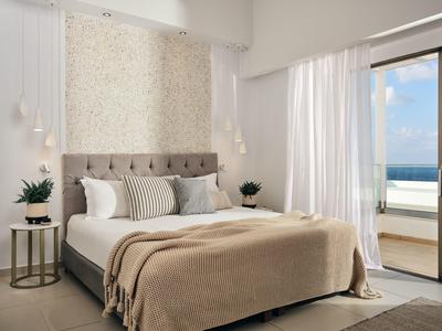 Helles Schlafzimmer mit großem Bett, beigem Überwurf, Pflanzen und Blick auf das Meer durch Balkonfenster.