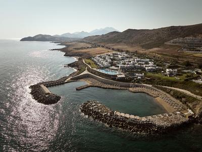 Kleiner Hafen mit Wellenbrechern an felsiger Küste, dahinter Siedlung und Hügel bei hellem Tageslicht.