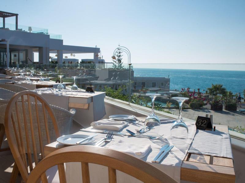 Terrassenrestaurant mit Holztischen und Stühlen, Blick auf Meer und Hafen im Hintergrund.