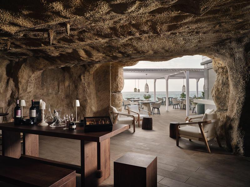 Innenraum einer gemütlichen, höhlenartigen Bar mit Holzmöbeln und Blick auf Außenterrasse bei Tageslicht.