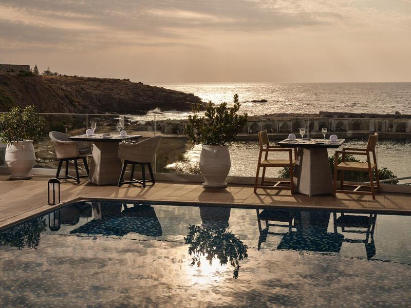 Terrasse mit Pool, Tischen, Stühlen und Pflanzen, Sonnenuntergang über Meer im Hintergrund.