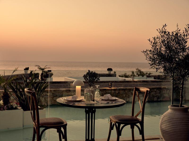 Tisch mit Kerze und Gläsern auf Terrasse, im Hintergrund ruhiges Meer bei Sonnenuntergang.