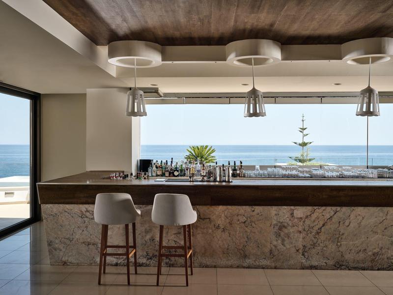 Moderner Barbereich mit Marmorfront, weißen Barhockern und Meerblick durch große Fenster.