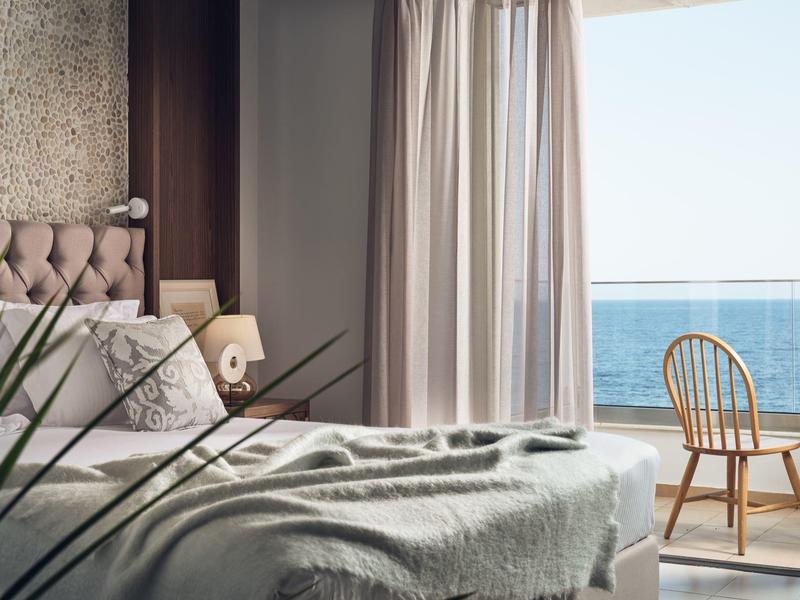 Gemütliches Schlafzimmer mit Bett, sanften Vorhängen und Holzstuhl vor Meerblick.