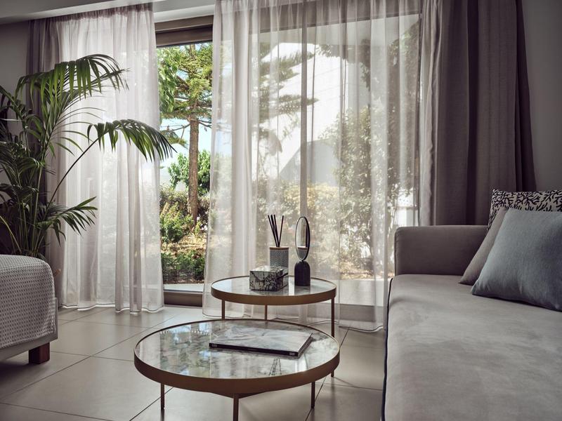 Helles Wohnzimmer mit grauem Sofa, zwei runden Glastischen, Pflanzen und großem Fenster mit Gardinen.