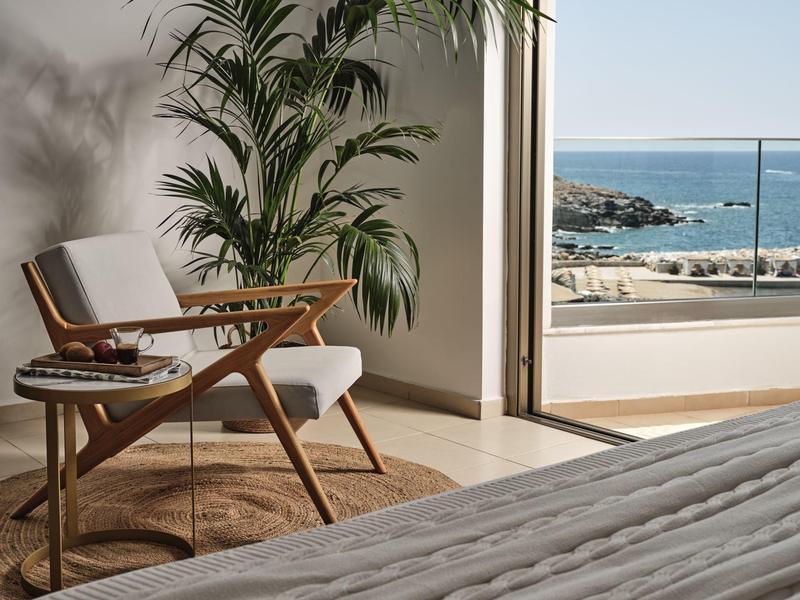 Hell eingerichtetes Zimmer mit Holzstuhl, Rundteppich, großer Pflanze und Blick auf das Meer.