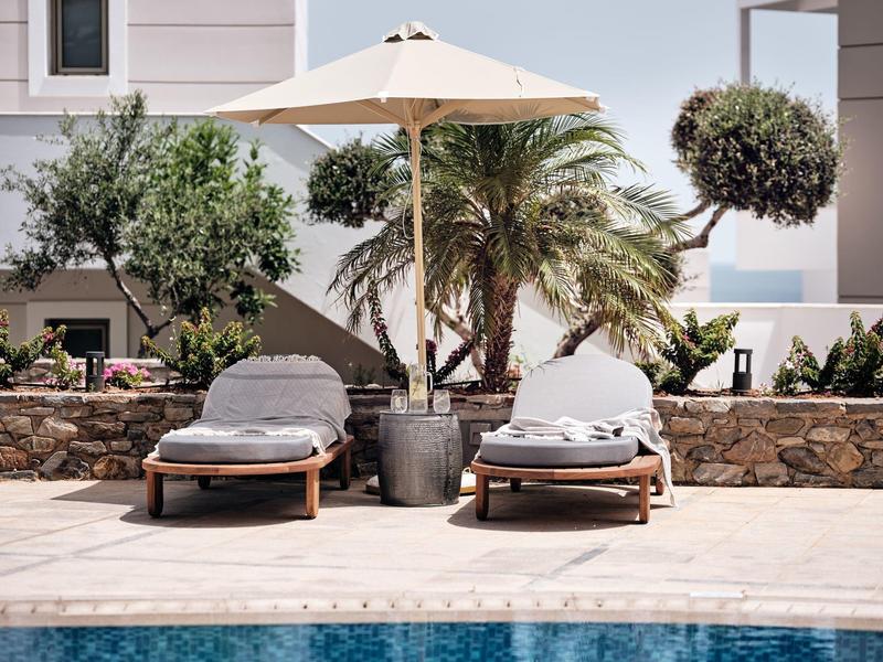 Zwei Holzliegestühle mit grauen Kissen unter weißem Sonnenschirm vor Palmen und Mauer.