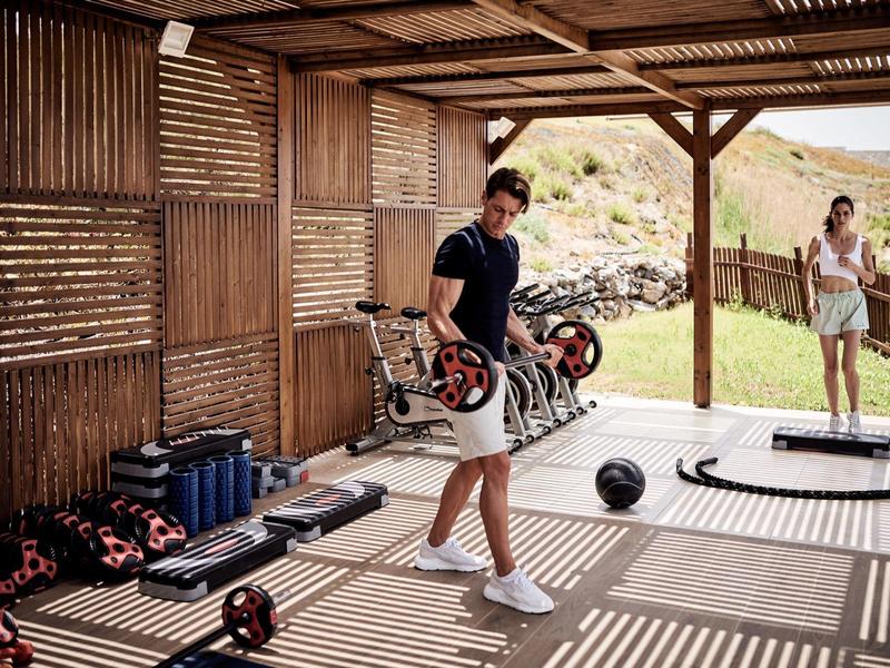 Mann hebt Hantel in offenem Holz-Sportbereich, Frau steht draußen auf Wiese, Gerätschaften liegen verteilt.