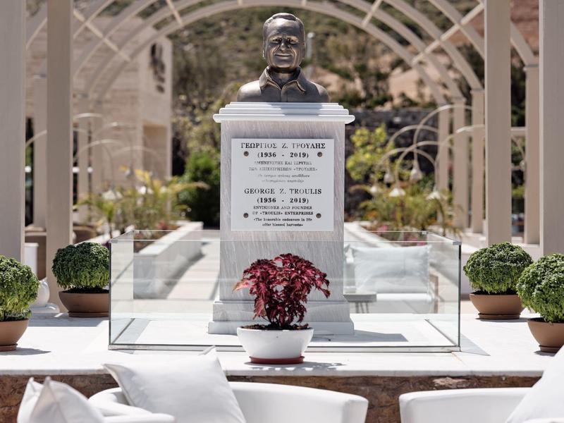 Moderner Gedenkplatz mit Büste, Glasumrandung, Pflanzen und weißen Sitzmöbeln vor felsiger Landschaft.