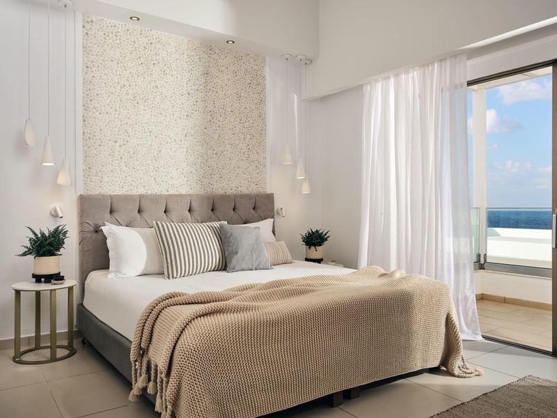 Helles Schlafzimmer mit großem Bett, beigem Überwurf, Pflanzen und Blick auf das Meer durch Balkonfenster.