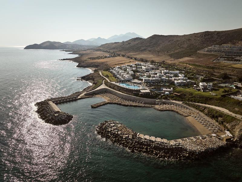 Kleiner Hafen mit Wellenbrechern an felsiger Küste, dahinter Siedlung und Hügel bei hellem Tageslicht.