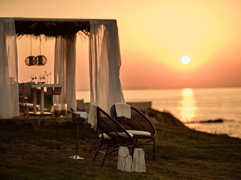 Zwei Stühle vor einer überdachten Lounge am Meer bei Sonnenuntergang mit goldgelbem Himmel.