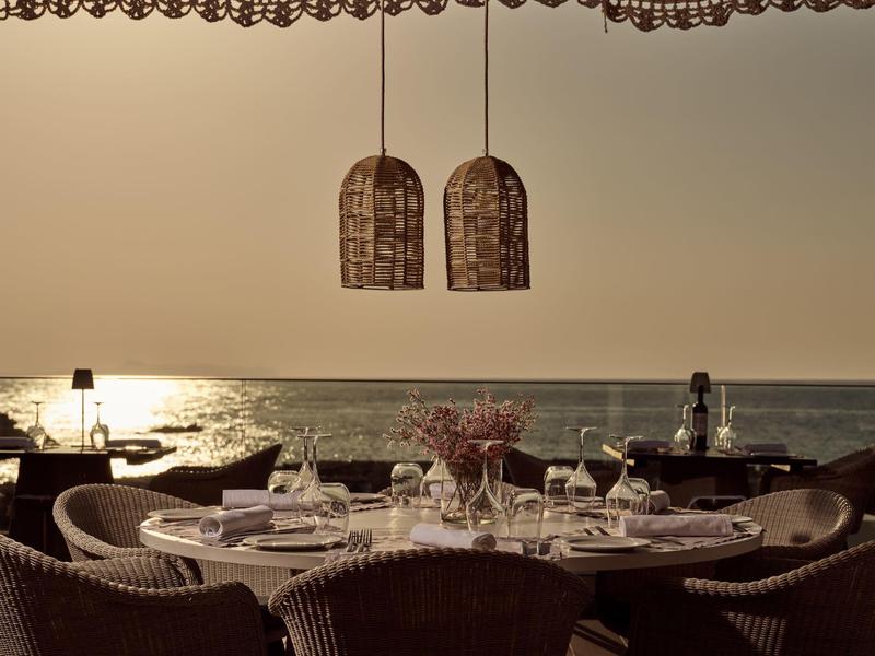 Gemütlicher Außenbereich mit gedecktem Tisch, Korbstühlen und Blick auf das Meer bei Sonnenuntergang.