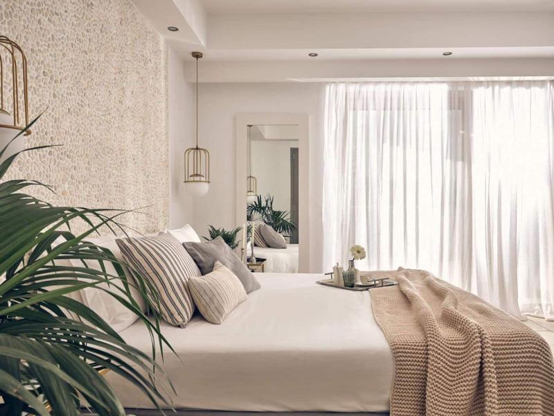 Modernes Schlafzimmer mit großem Bett, Kissen, Decke und Pflanzen neben dem Fenster mit Vorhängen