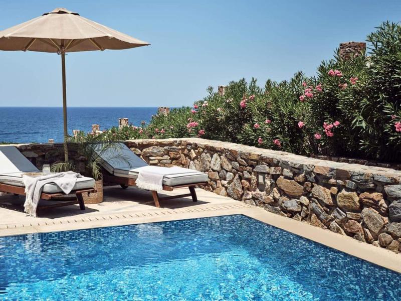 Swimmingpool mit Liegestühlen, Sonnenschirm und Meerblick, umgeben von Steinmauer und Blumen.