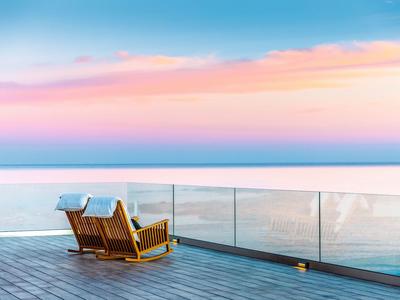 Zwei Holzliegen auf einer Terrasse mit Glasgeländer mit Blick auf einen ruhigen, bunten Himmel über dem Meer.