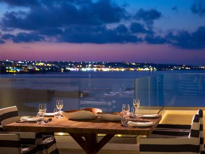 Terrasse avec table en bois dressée offrant une vue sur une ville côtière illuminée au coucher du soleil.