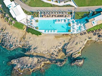 Luftaufnahme eines Hotels mit Pool neben einem felsigen Strand und klarem Meerwasser.