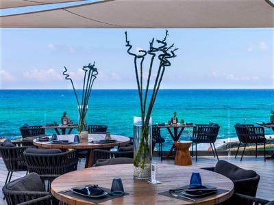 Terrasse mit Tischen und Stühlen mit Blick auf das blaue Meer unter einem Sonnensegel.