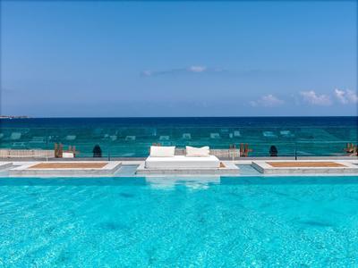 Infinity-Pool mit klar blauem Wasser, weißen Liegen und ruhigem Meer im Hintergrund unter blauem Himmel.