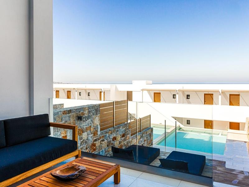 Balkon met zitgedeelte met uitzicht op een zwembad en witte gebouwen onder een heldere lucht.