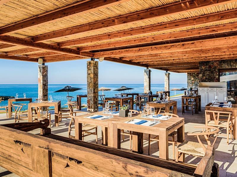 Openluchtrestaurant met houten meubilair en zeezicht onder een rustiek houten dak.