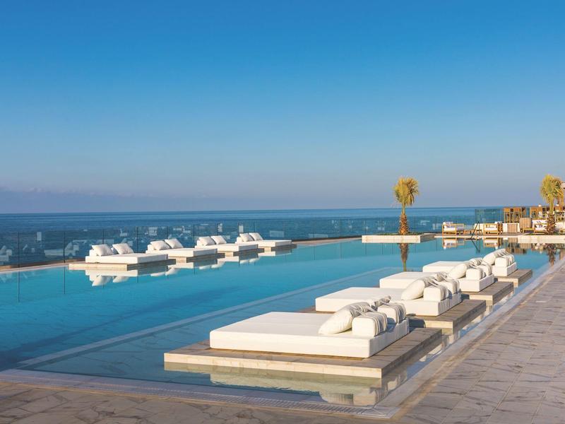 Infinity zwembad met uitzicht op de oceaan, witte ligbedden en een heldere blauwe lucht.