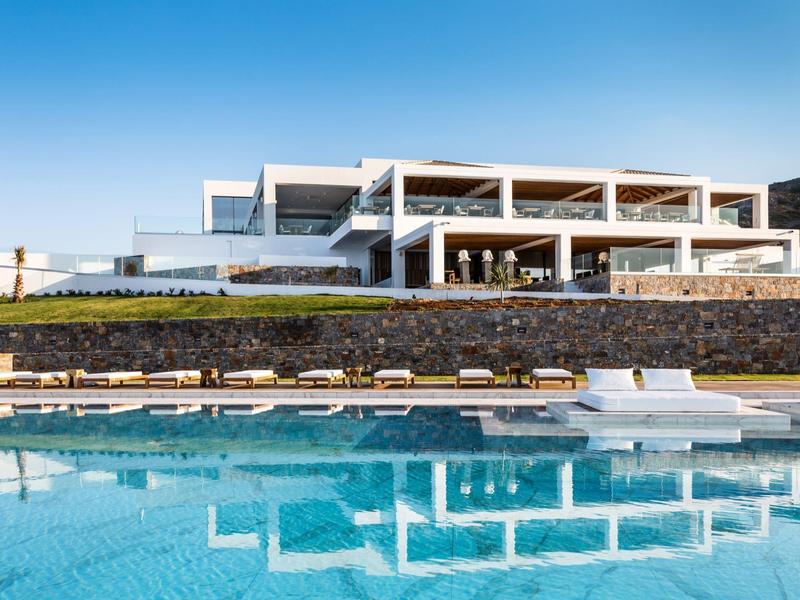 Modern hotel met groot zwembad en ligbedden onder een heldere blauwe lucht.